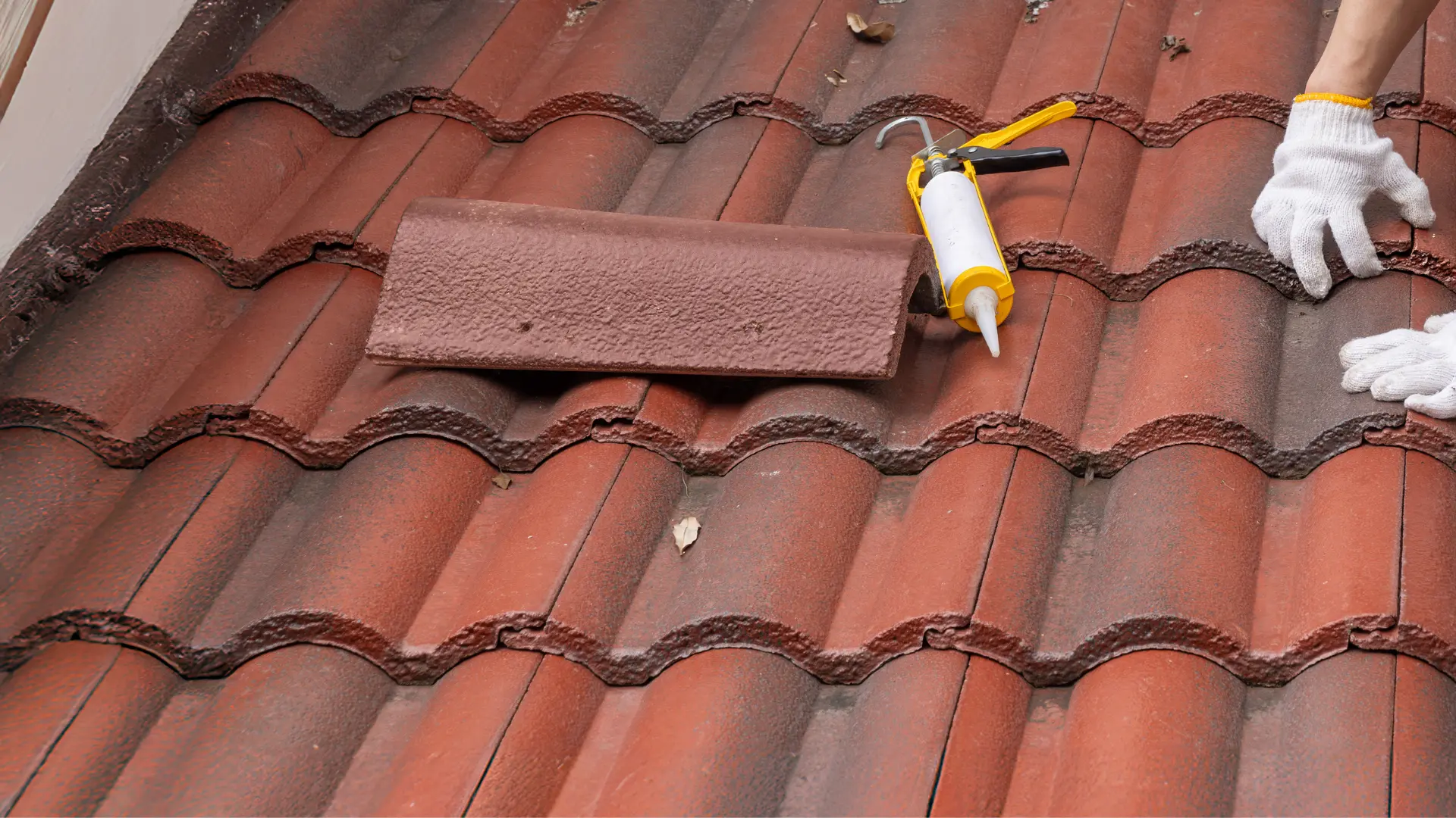 Roofer in gloves replacing a ridge tile with sealant gun on a red clay tiled roof
