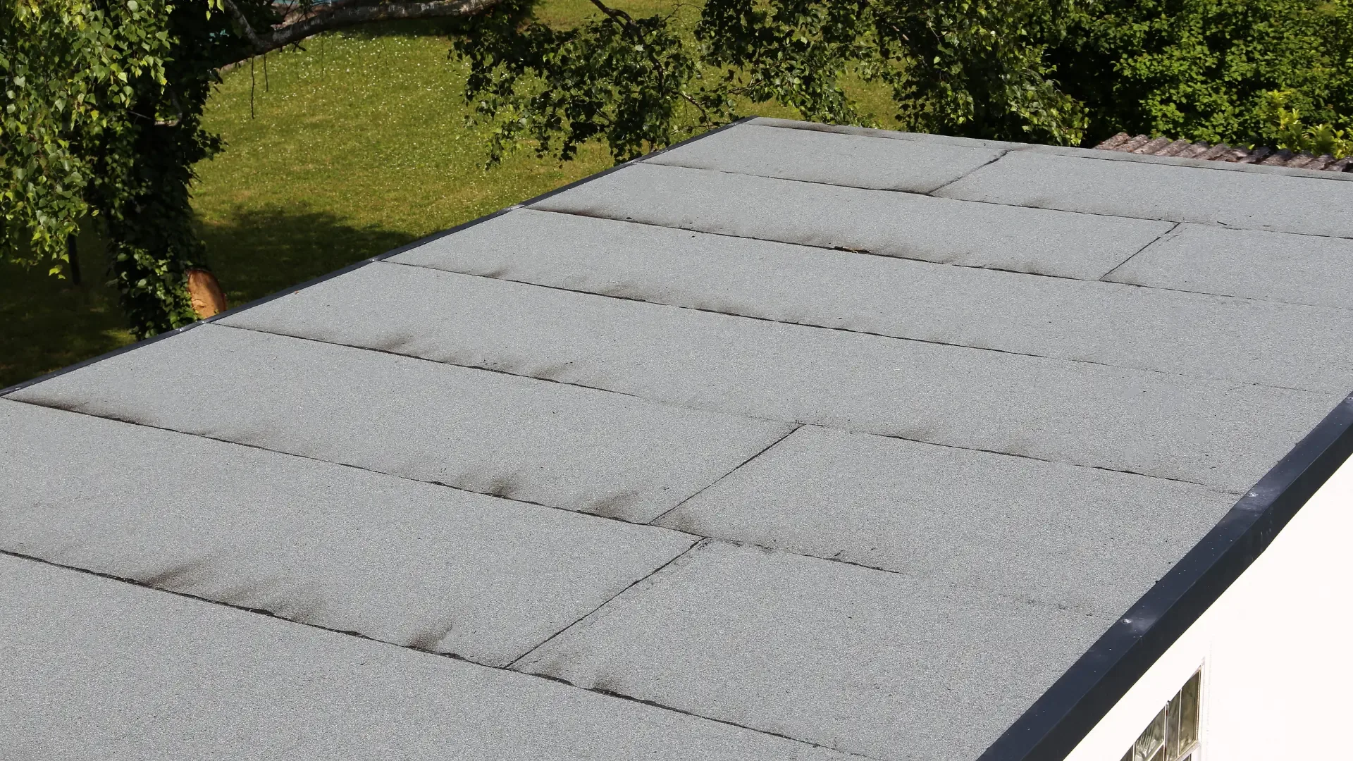 Felt flat roof with overlapping mineral-finish sheets and a dark edge trim on a white-walled extension